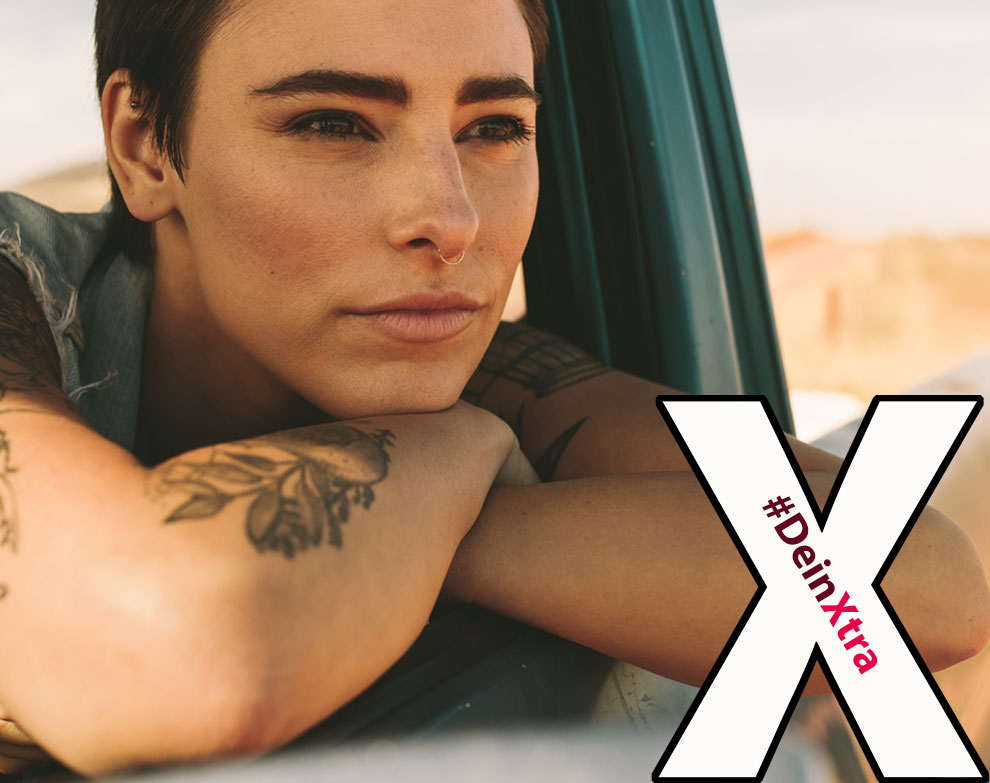 © AdobeStock: Jacob Lund #DeinXtra | Bild zeigt eine mittelalte Frau im Auto mit Tattoos, die in die Ferne schaut. | © AdobeStock: Jacob Lund